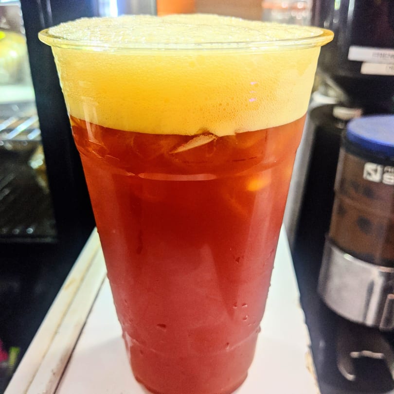 Close-up of a vibrant red drink with a thick layer of yellow foam on top, in a clear plastic cup.