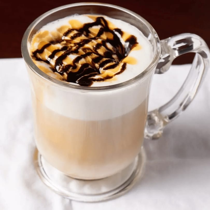 A clear mug of latte with foamed milk, drizzled with chocolate and caramel syrup.