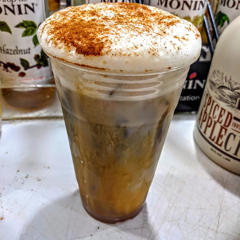 Iced coffee with a thick layer of foam and cinnamon powder on top, with syrup bottles in the background.